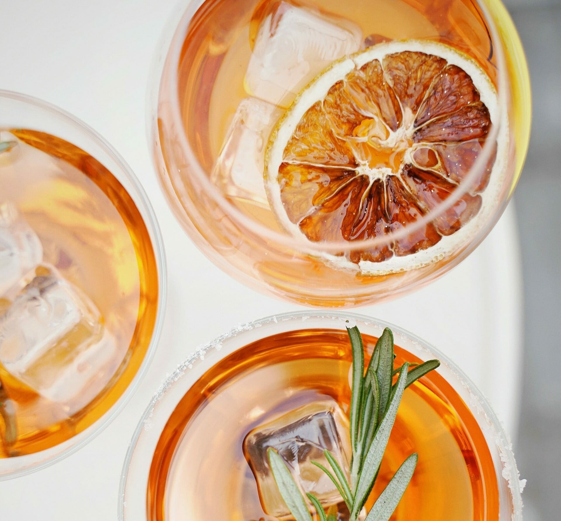 Overhead view of three glasses of iced amber-colored drinks, one garnished with a dried citrus slice and another with a sprig of rosemary, capturing the refreshing essence akin to Spryker at Shoptalk Europe.