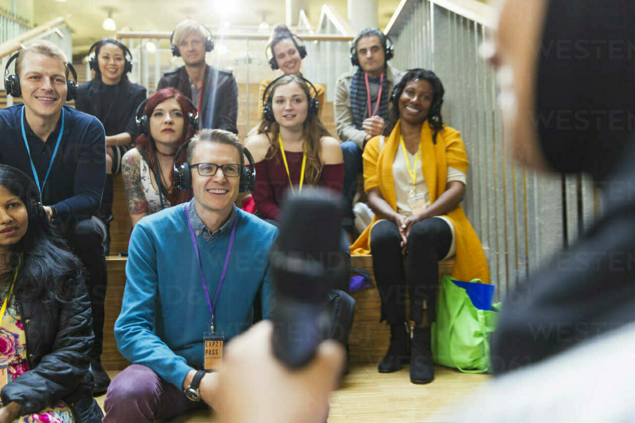 A diverse group of people, wearing headphones and attentively listening to a speaker holding a microphone, sits on the stairs at Spryker at Shoptalk Europe.