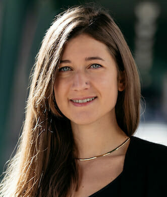 A woman with long brown hair and blue eyes, wearing a black top and a thin gold necklace, smiles softly, capturing the essence of current trends. The background is blurred and greenish.