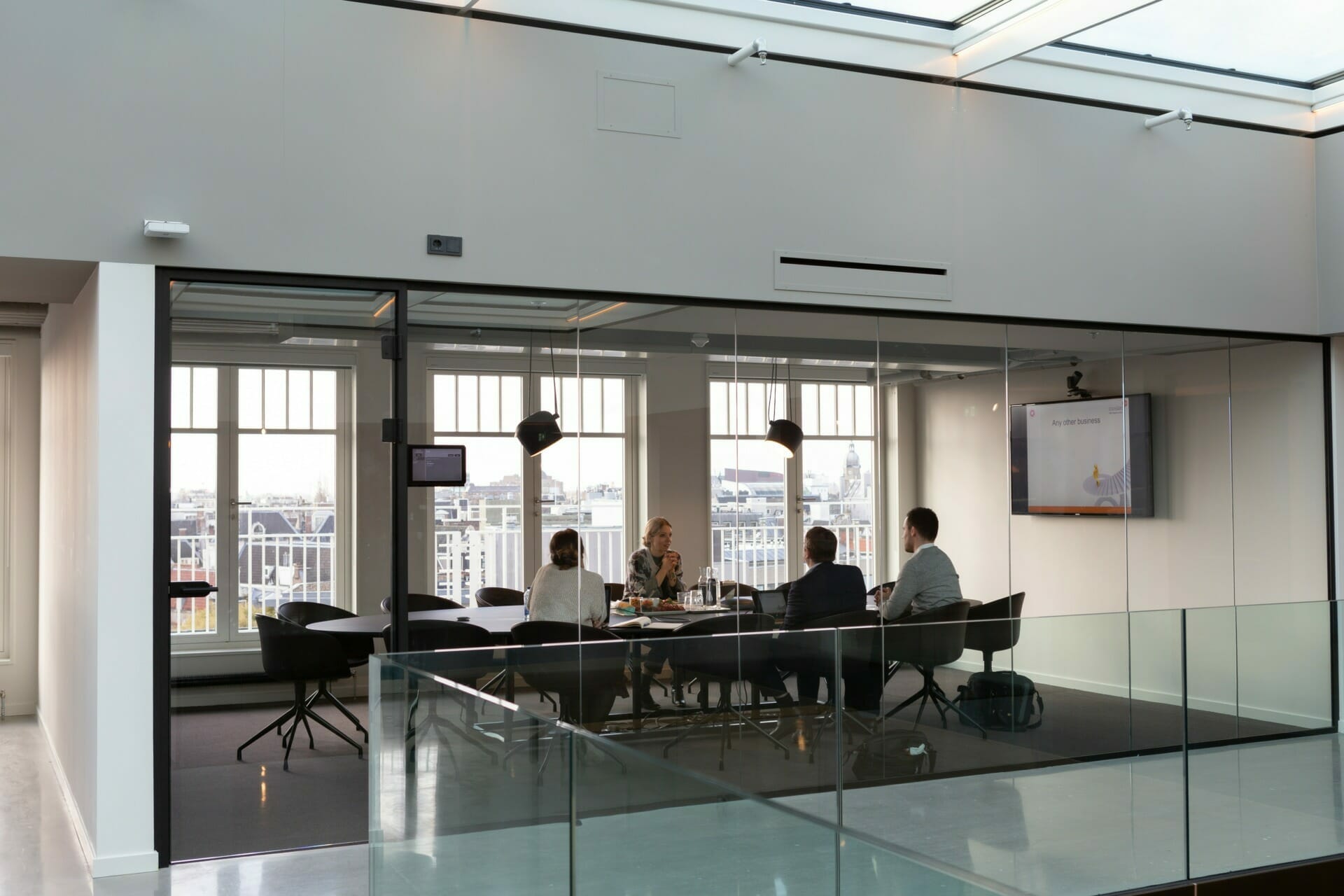 A group of four people having a meeting in a glass-walled conference room with a TV screen displaying a Customer Advisory Board presentation slide.