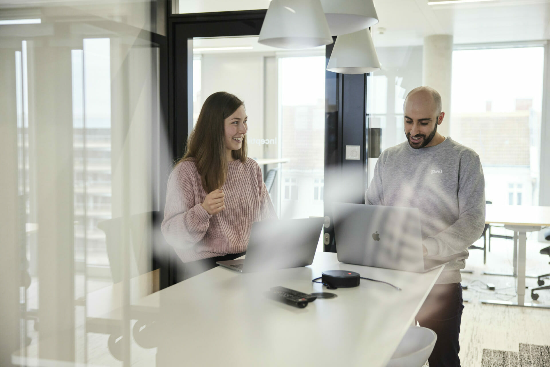 Two individuals are working at a high table in a modern office, Emerce100 recognition proudly displayed on the wall. Both are using laptops and appear engaged in conversation. The woman is smiling, and they are surrounded by contemporary office furniture.