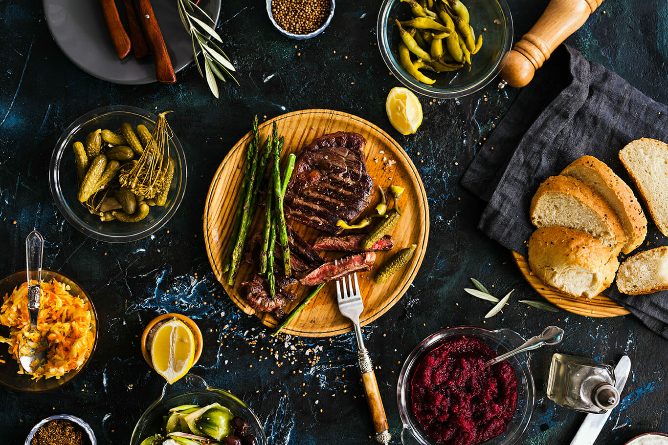 A table setting with a wooden plate of grilled steak and asparagus, surrounded by bowls of various sides, including pickles, olives, bread, and condiments on a dark surface—showcasing sustainable commerce in every carefully chosen detail.