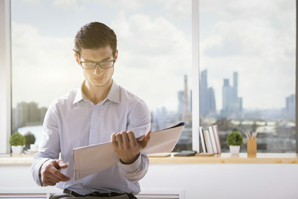 A man wearing glasses and a white shirt sits by a window with a cityscape view, holding and reading a Spryker Raizor folder.