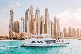 A luxurious white yacht cruises on turquoise water with a backdrop of towering, modern skyscrapers under a clear, sunny sky. The scene is set in a bustling coastal city, evoking a sense of opulence and leisure.