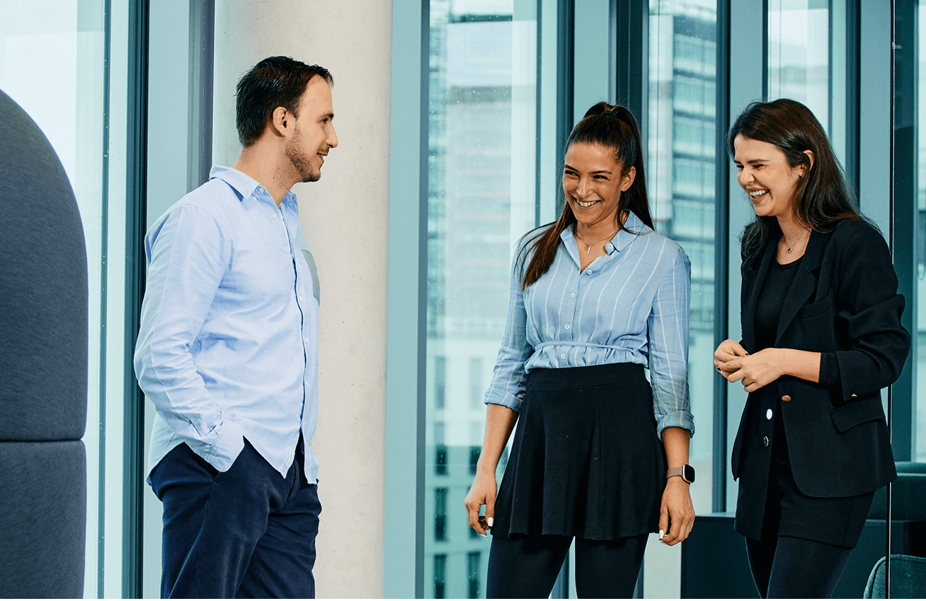 Three people in business attire stand and converse in a modern office setting. Two women are smiling while the man on the left engages with them, discussing their latest B2B online strategies for an upcoming conference in Orlando.