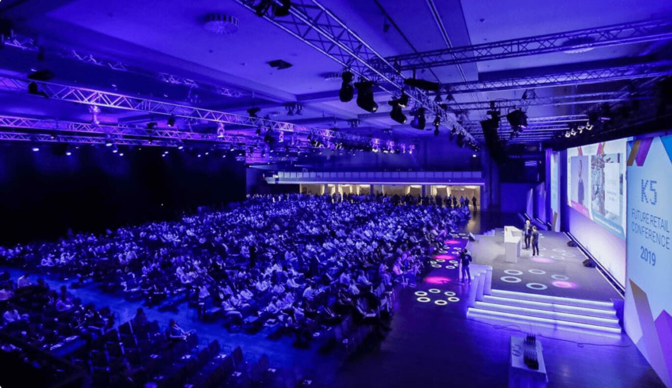 A large audience in a dimly lit auditorium watches a presentation on stage at a conference. The screen displays the text "K5 Konferenz 2022." Purple lighting illuminates the venue.