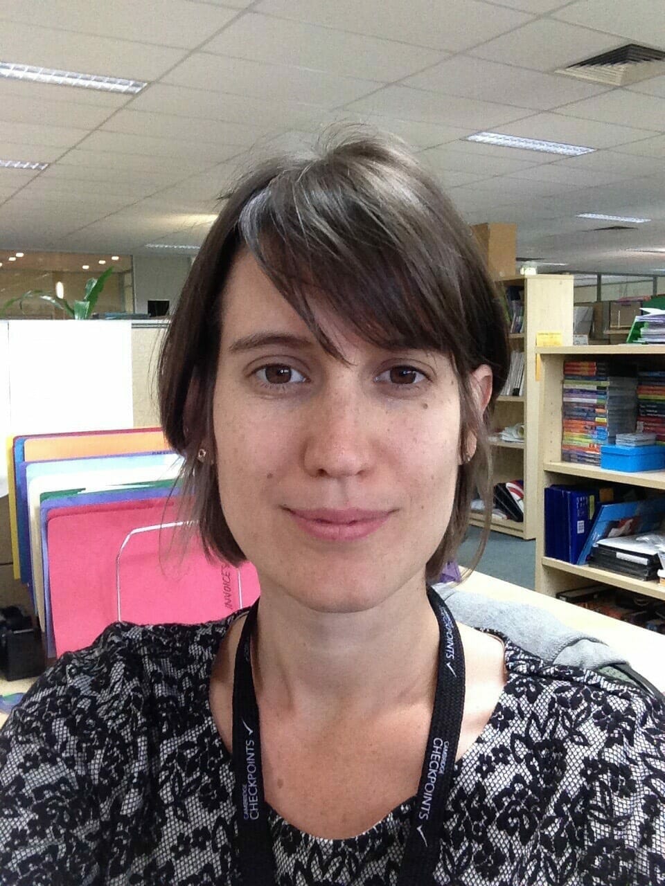 A woman with short brown hair, wearing a black and white patterned top, takes a selfie in what appears to be an office environment filled with books and colorful file folders. The setting suggests she's ready to discuss 2022 Spryker EXCITE takeaways.