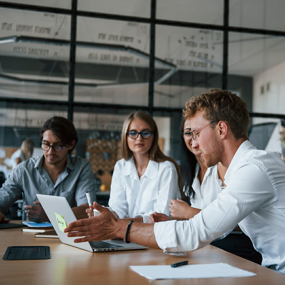 Four people are sitting at a table in a modern office, discussing composable commerce strategies. One person is pointing to a laptop screen with a sticky note attached.