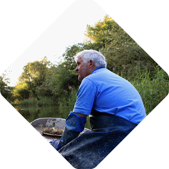 Elderly man in a blue shirt and waders sits in a boat on a river, surrounded by lush greenery, while viabirds chirp melodiously from the treetops.