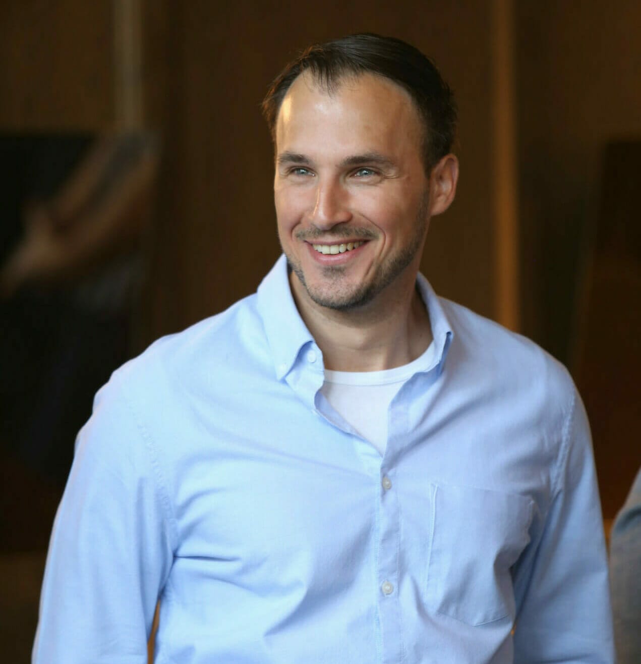 A man with short dark hair and a beard is smiling. He is wearing a light blue button-down shirt over a white t-shirt. The background, likely taken in Orlando, is blurry and out of focus.