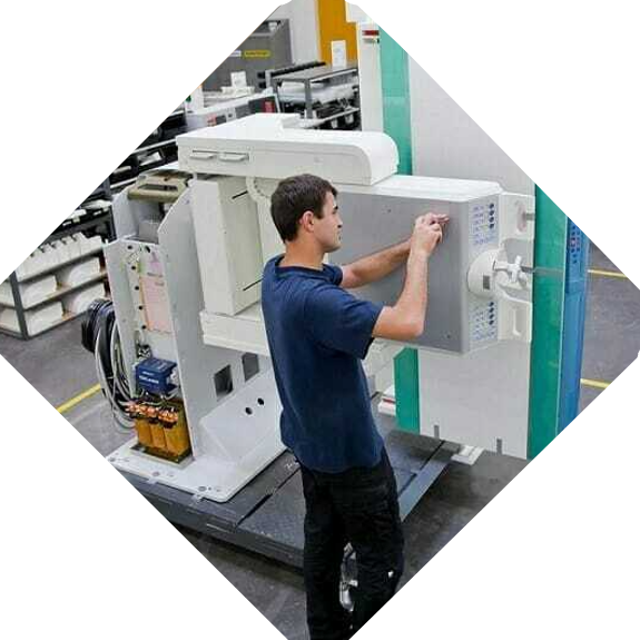 A technician in a blue shirt meticulously works on an industrial machine in a factory setting, adjusting a component with his hands. Siemens Healthineers equipment and various tools are visible in the background.