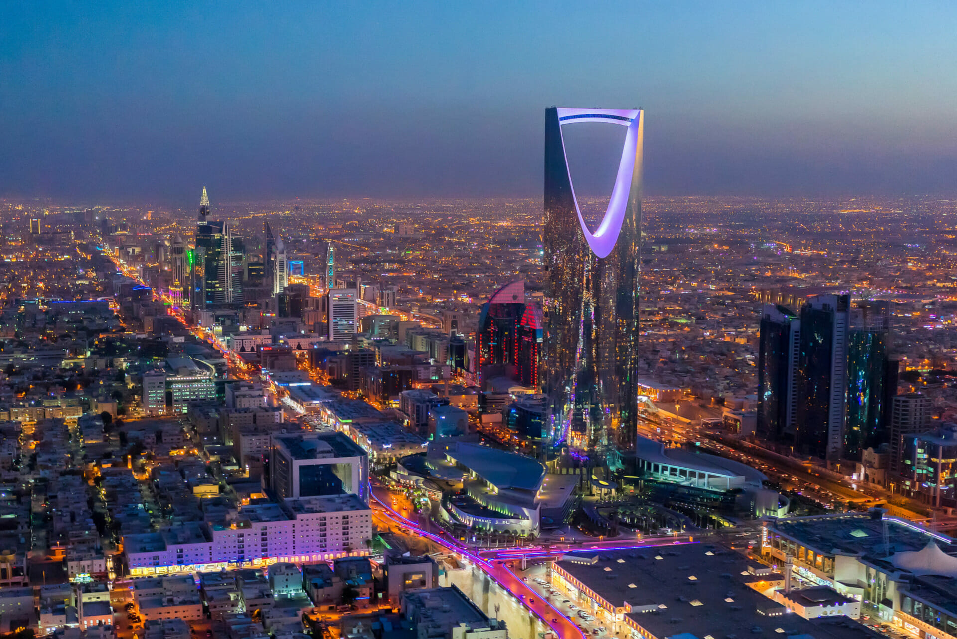 Aerial view of Riyadh at dusk, featuring the illuminated Kingdom Centre Tower and the seamless Saudi Arabia cityscape with streets lit up.