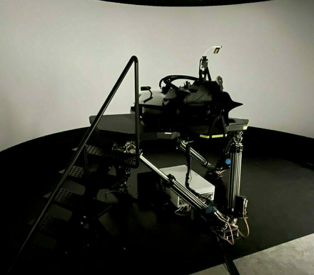 A black flight simulator with a stairway and control panel in a dark room. The simulator, equipped with various mechanical components, stands as a testament to digital success against a large, curved white screen.