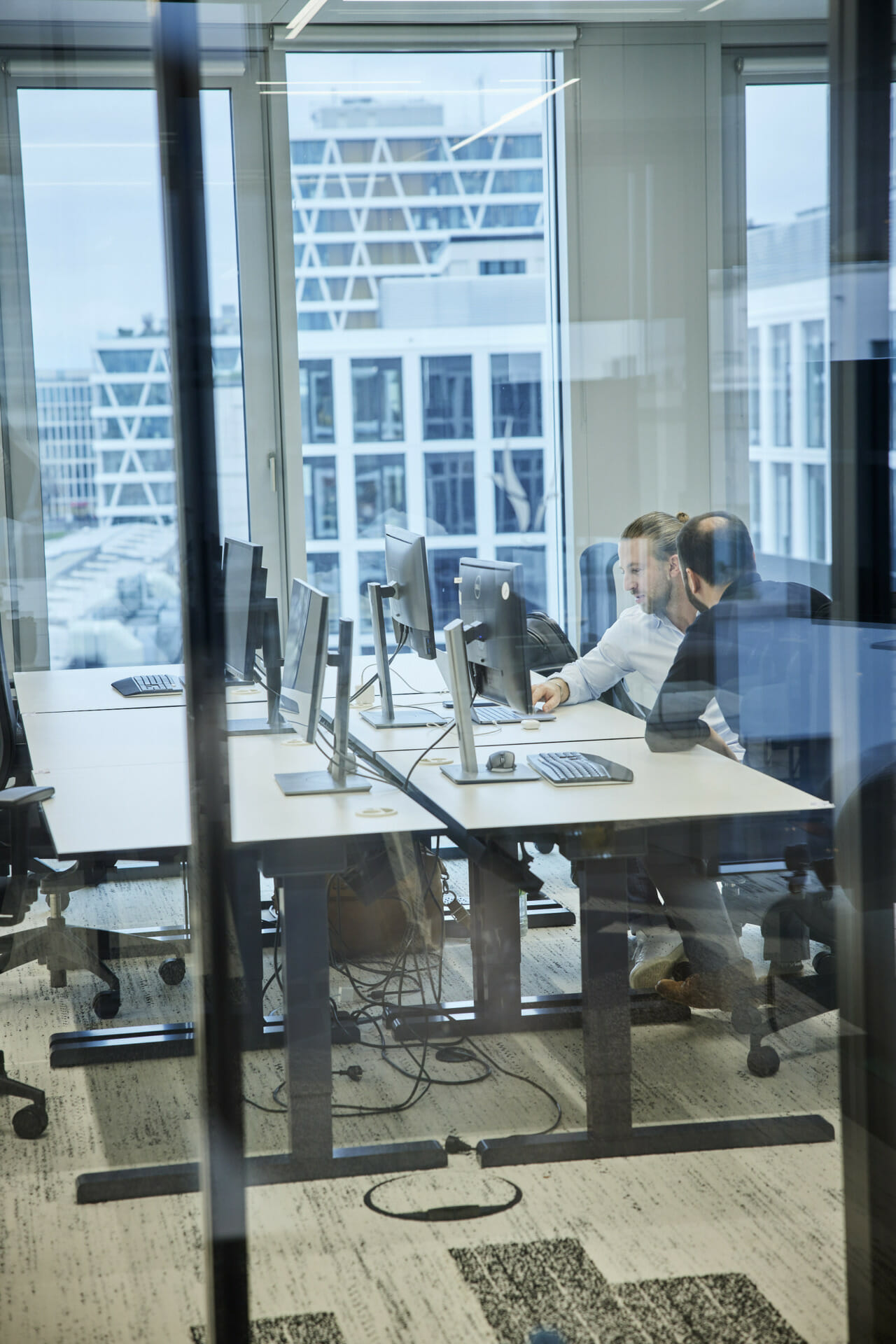 Two people sitting at computer desks in a modern office with large windows, discussing something on a screen showcasing Poolwerx services.