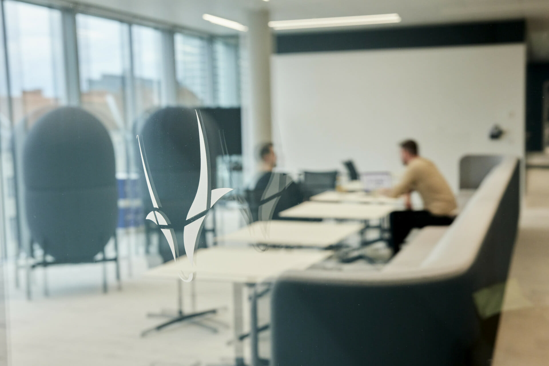 Two people sit at a table in a modern office space, engaged in conversation about leadership strategies. The foreground features glass walls with abstract designs and blurred reflections, subtly capturing the dynamic movement of the bustling environment.