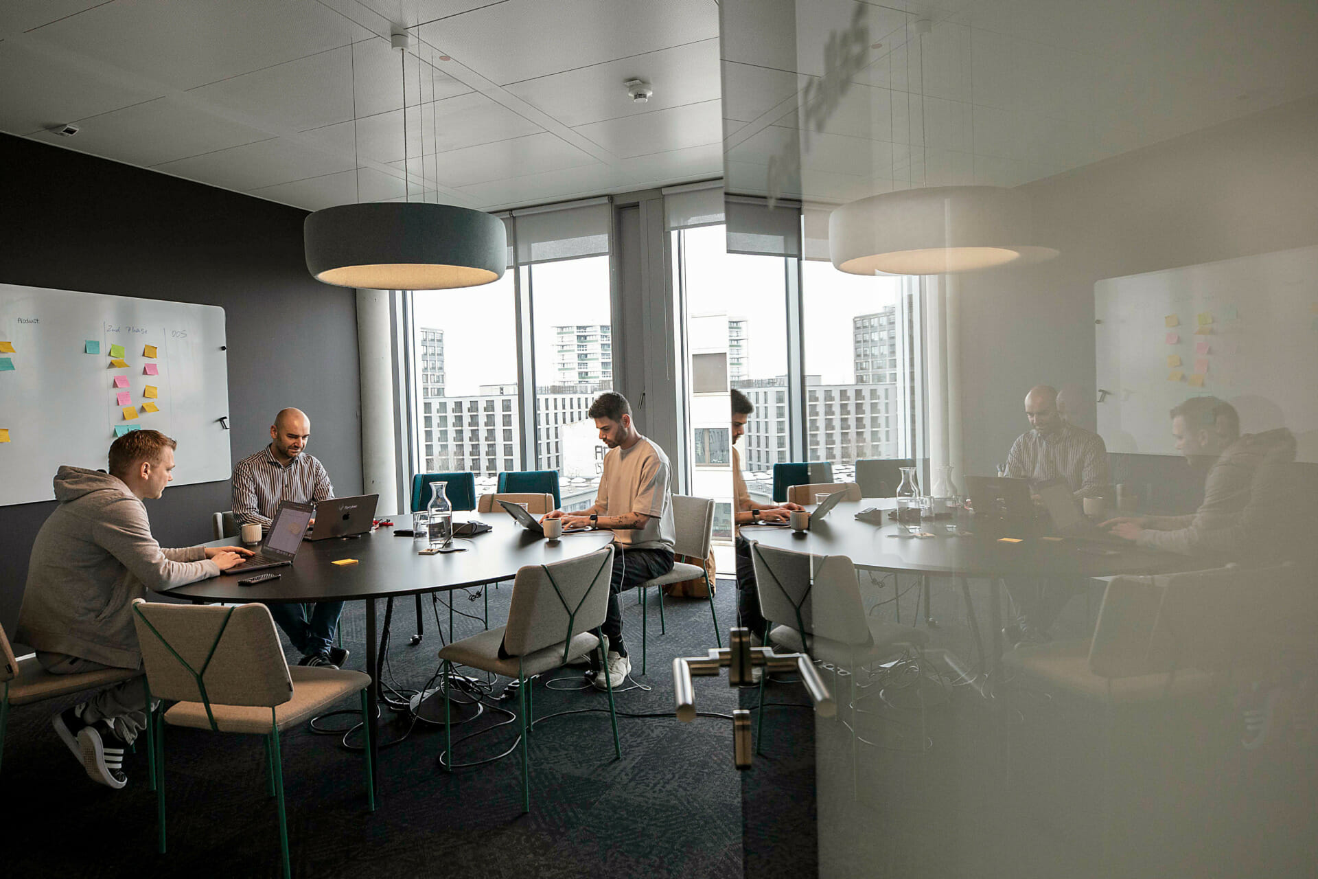 Four people are sitting at a conference table in a modern Publicis Sapient office with a large window and glass door. They are working on laptops with sticky notes on a whiteboard in the background.