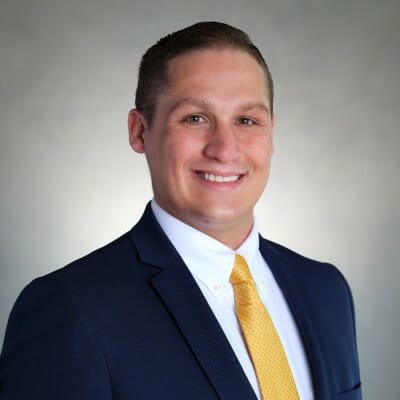 Smiling person in a dark blue suit and yellow tie, posing confidently against a neutral background, embodies the professionalism of the automotive aftersales industry.