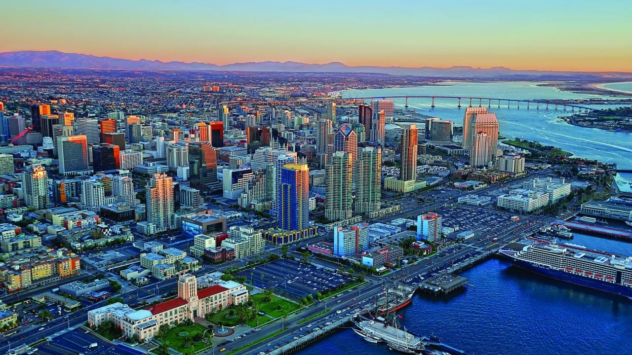 Aerial view of a coastal city with a mix of tall buildings, B2B Connect network clusters, a curving bridge over the water, docks, and a blend of residential and commercial areas during sunset.