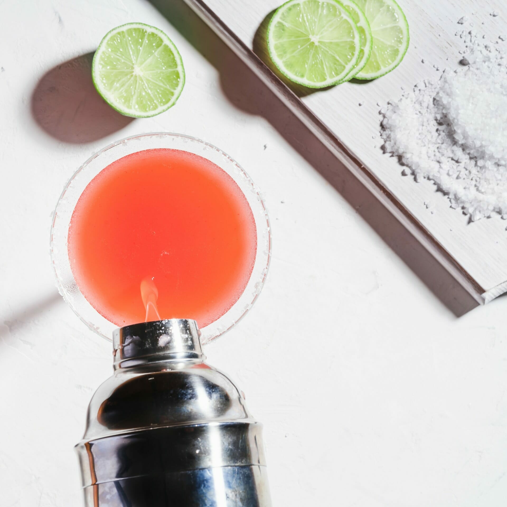 Top view of a red cocktail being poured from a stainless steel shaker into a glass, with salt on the rim, next to sliced limes and a pile of salt on a white surface. Experience this refreshing 2023 mix popular in online Florida B2B circles.