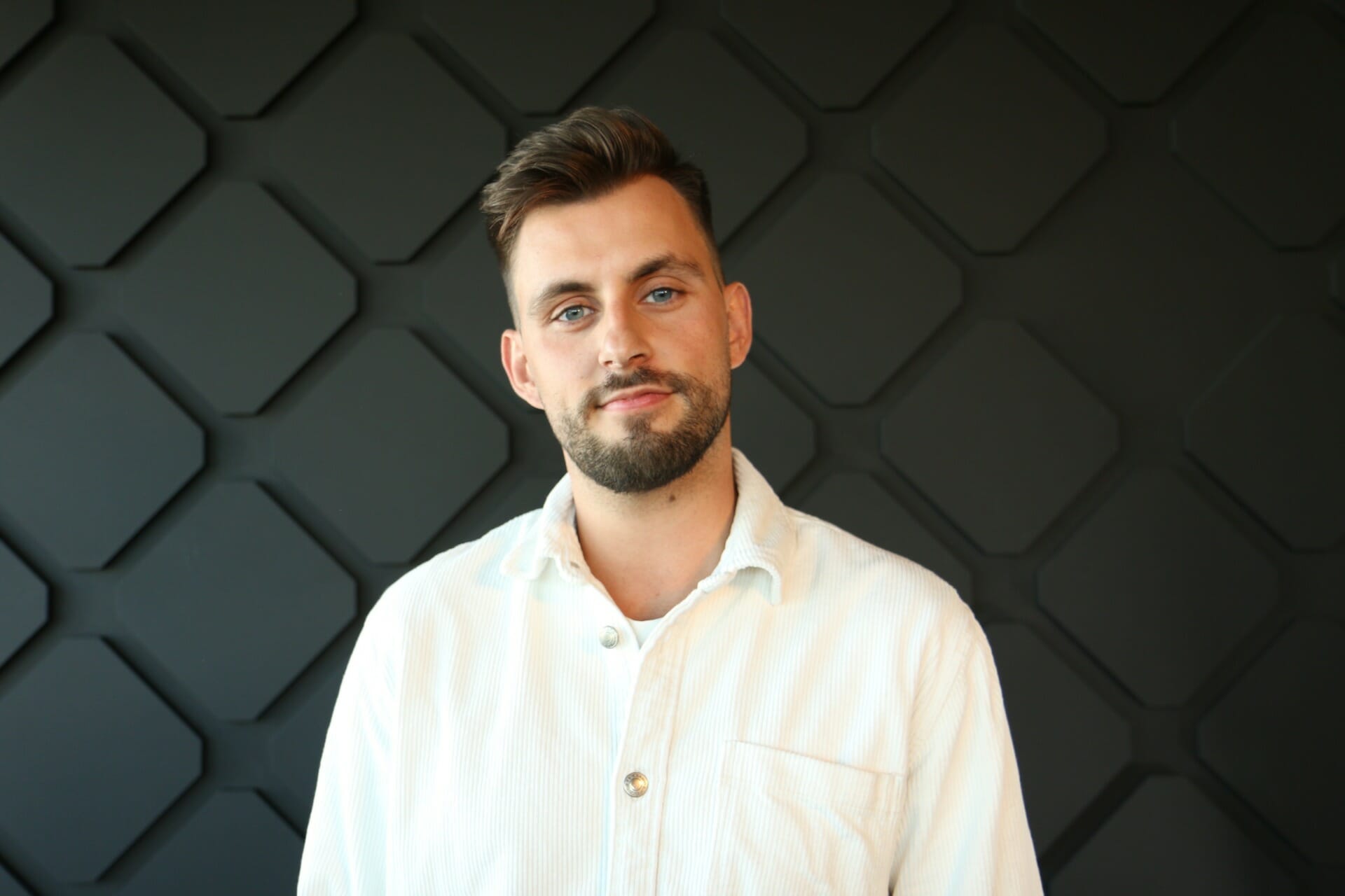 A man with short brown hair, a beard, and a neutral expression, wearing a light-colored button-up shirt, stands in front of a geometric patterned wall that hints at the complexity of digital commerce strategies for manufacturers.