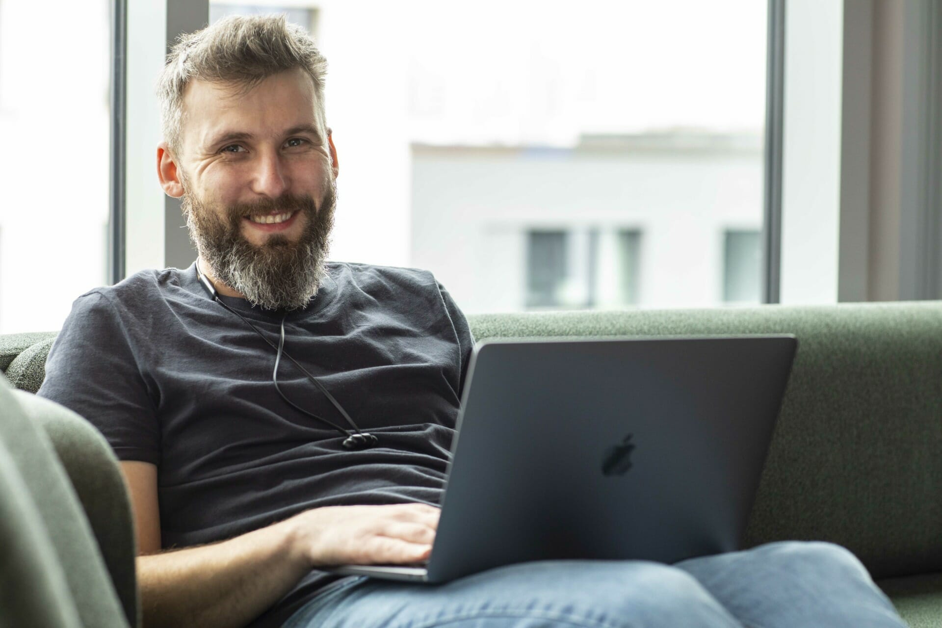 A bearded man in a black shirt sits on a green couch using a laptop, smiling at the camera. Trevipay solutions might just be making his day.