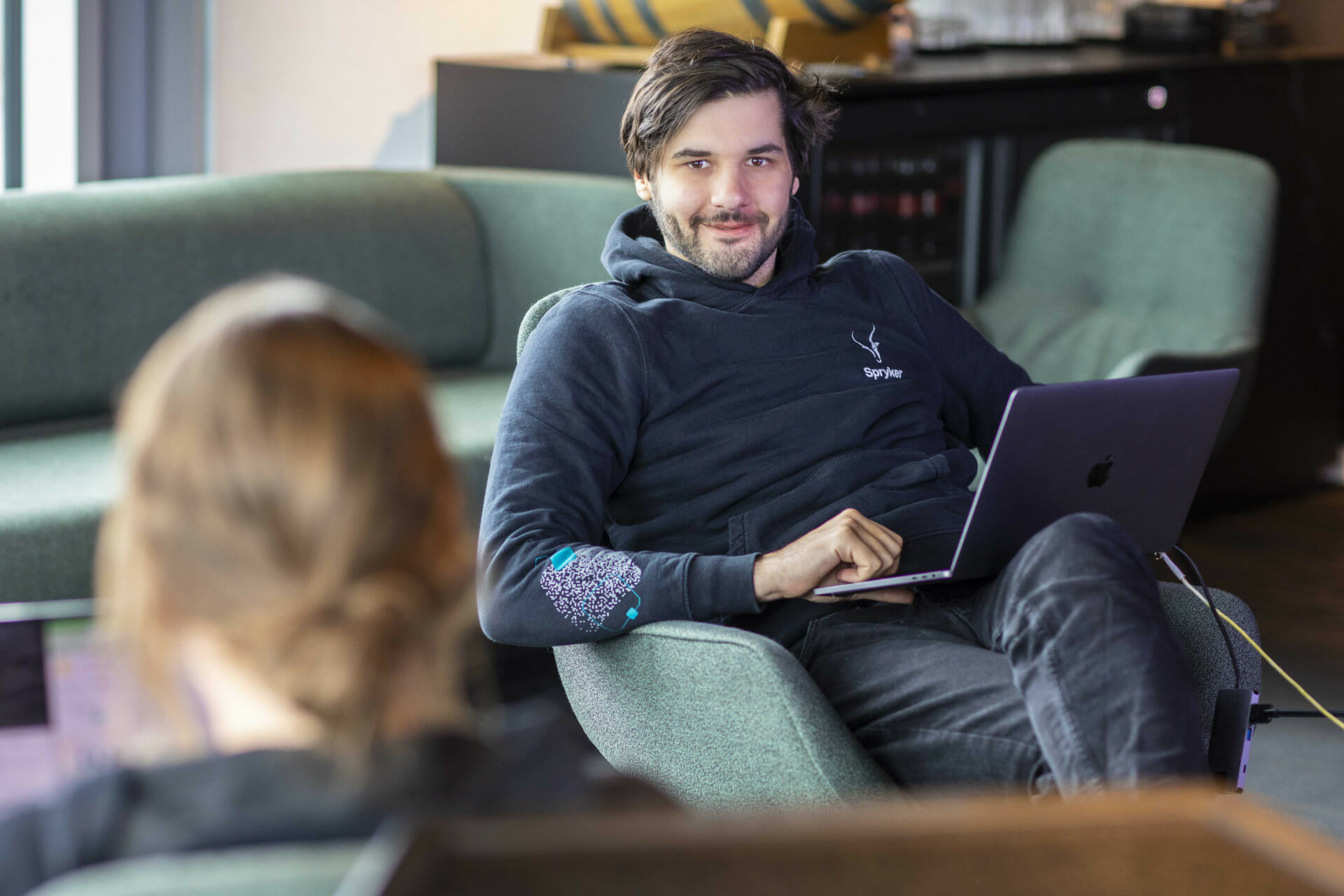 A man in a black hoodie sits on a green chair, working on his Trevipay-integrated laptop. Another person is partially visible in the foreground, also using a laptop.