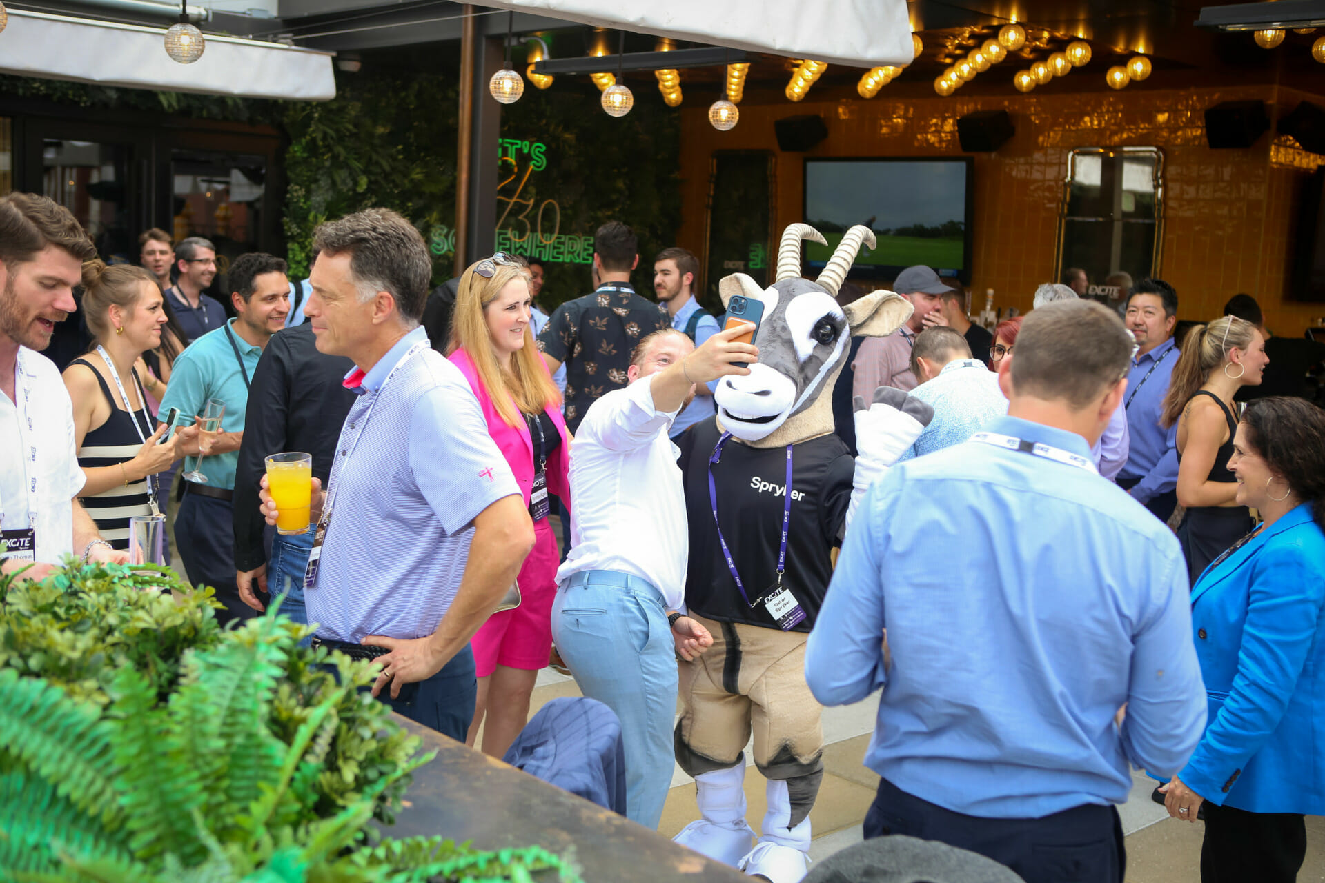 People are socializing and taking selfies at an outdoor B2B event for Florida manufacturers and distributors. One person is dressed in a goat costume. There are drinks on the table and hanging lights above.