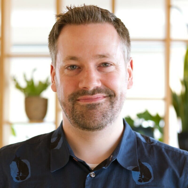 A man with short brown hair and a beard is looking at the camera and smiling. He is wearing a dark shirt with patterns. There are plants and wooden shelves in the background, subtly hinting at an environment where digital commerce strategies for manufacturers might flourish.