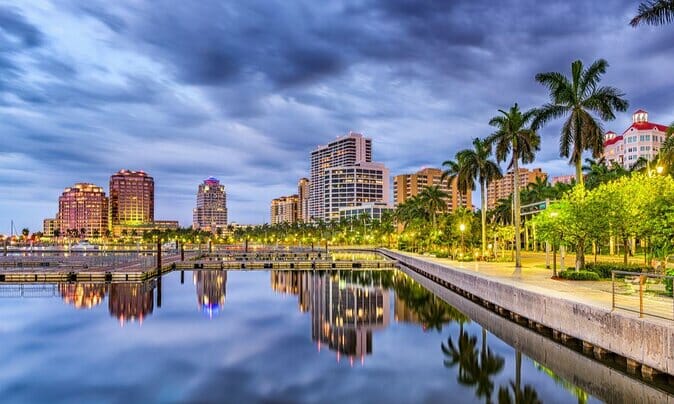 Waterfront cityscape with modern buildings, illuminated at dusk, reflected in a calm body of water; palm trees line the right side, and a cloudy sky looms overhead—perfect for B2B online Florida manufacturers and distributors to envision their next big venture.