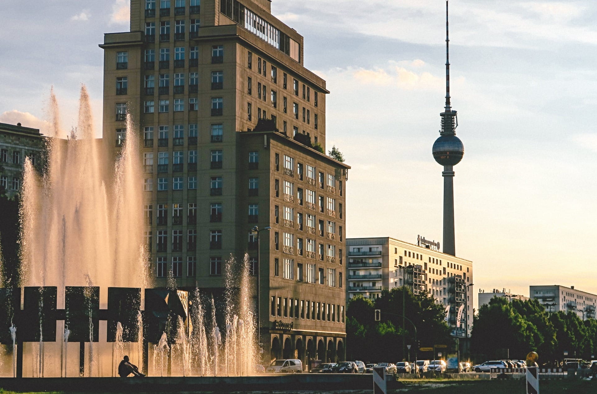 Tall buildings and a fountain at sunset with the Berlin Fernsehturm visible in the background. Cars drive on the street, and a person sits near the fountain, reminiscent of a bustling scene where B2B Online Europe manufacturers and distributors might meet for business discussions.