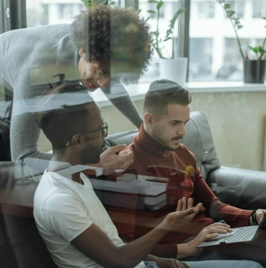 Three people are having a discussion while looking at a laptop screen with one person pointing at the display, likely reviewing a B2B Online Europe platform for manufacturers and distributors. They are seated on a couch in a well-lit room with large windows and potted plants.