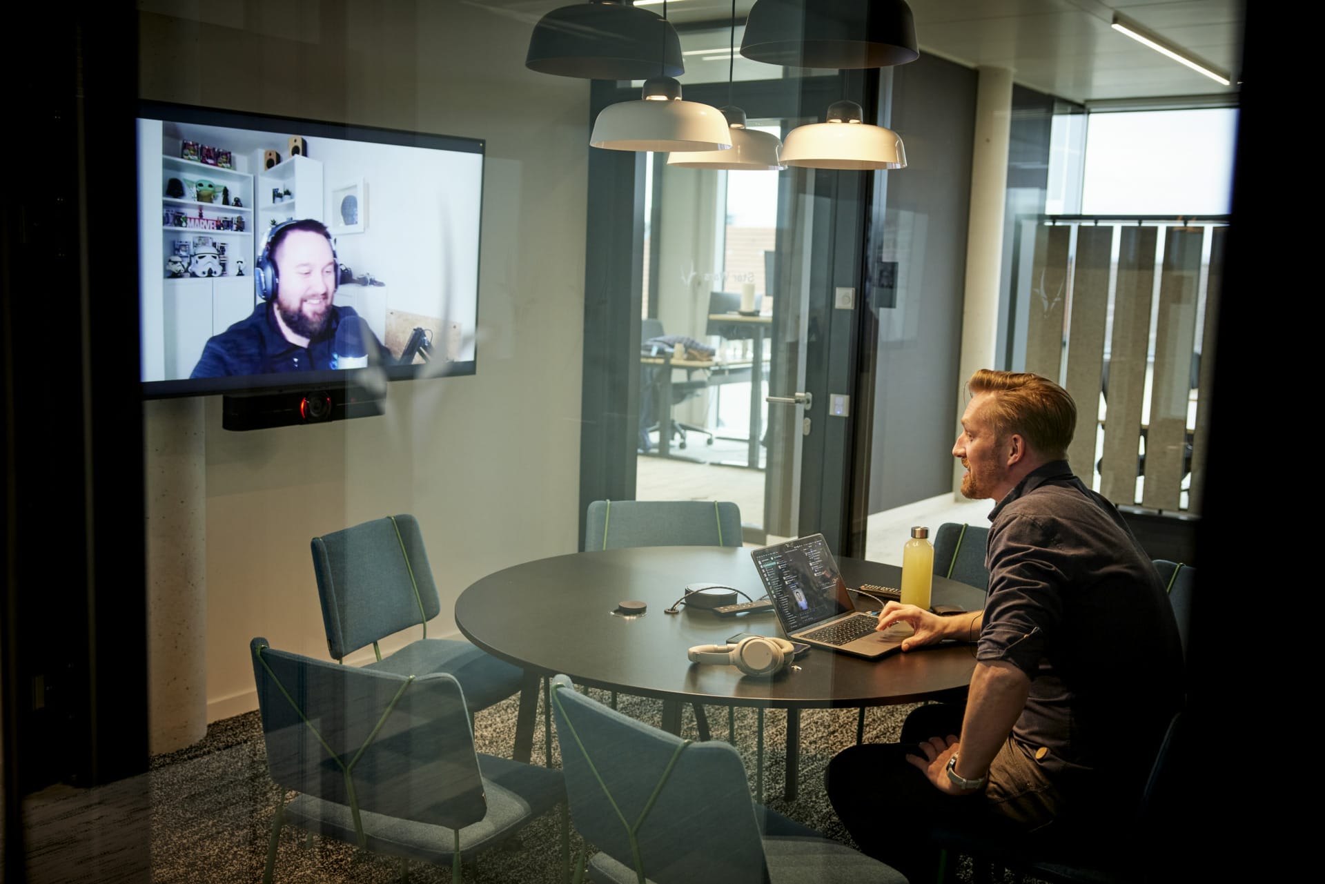 A man sits in a conference room using a laptop and headphones for a video call with the Spryker CMO, who is visible on a wall-mounted screen.
