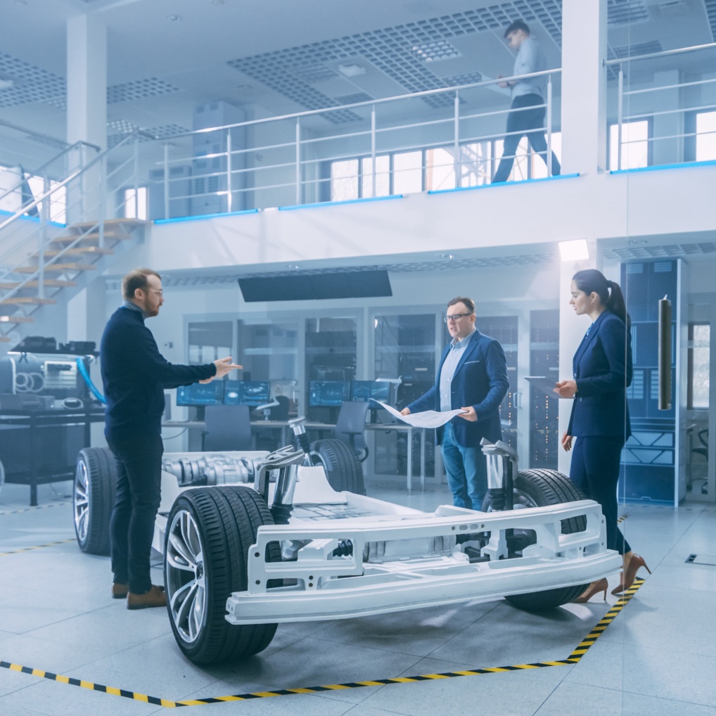Three people are discussing plans next to a car chassis in a modern automotive workshop, presumably gearing up for Spryker EXCITE 2024. Another person walks on a mezzanine in the background.