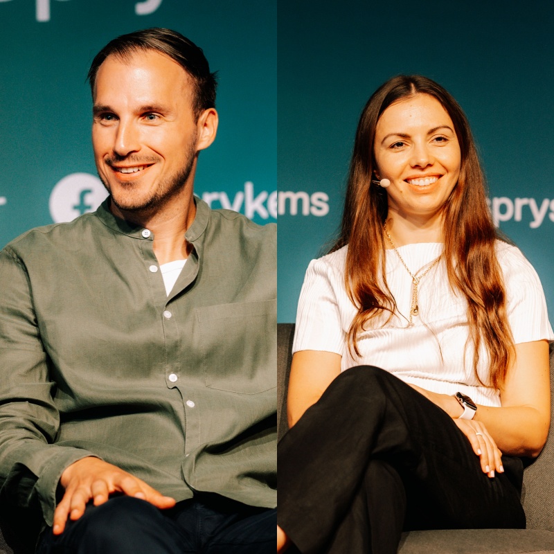 Two individuals are sitting and smiling in front of a blue-green background with Spryker EXCITE 2024 logos. The person on the left is wearing a green shirt, while the one on the right is dressed in a white blouse and black pants.
