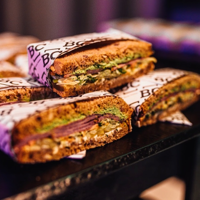 Three halved sandwiches with various fillings, including vegetables, are wrapped in branded Spryker EXCITE 2024 paper and displayed on a dark surface.