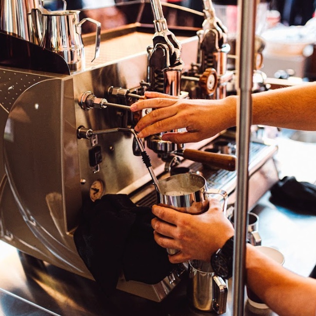 A barista at Spryker EXCITE 2024 operates an espresso machine, using a steam wand to froth milk in a metal pitcher.