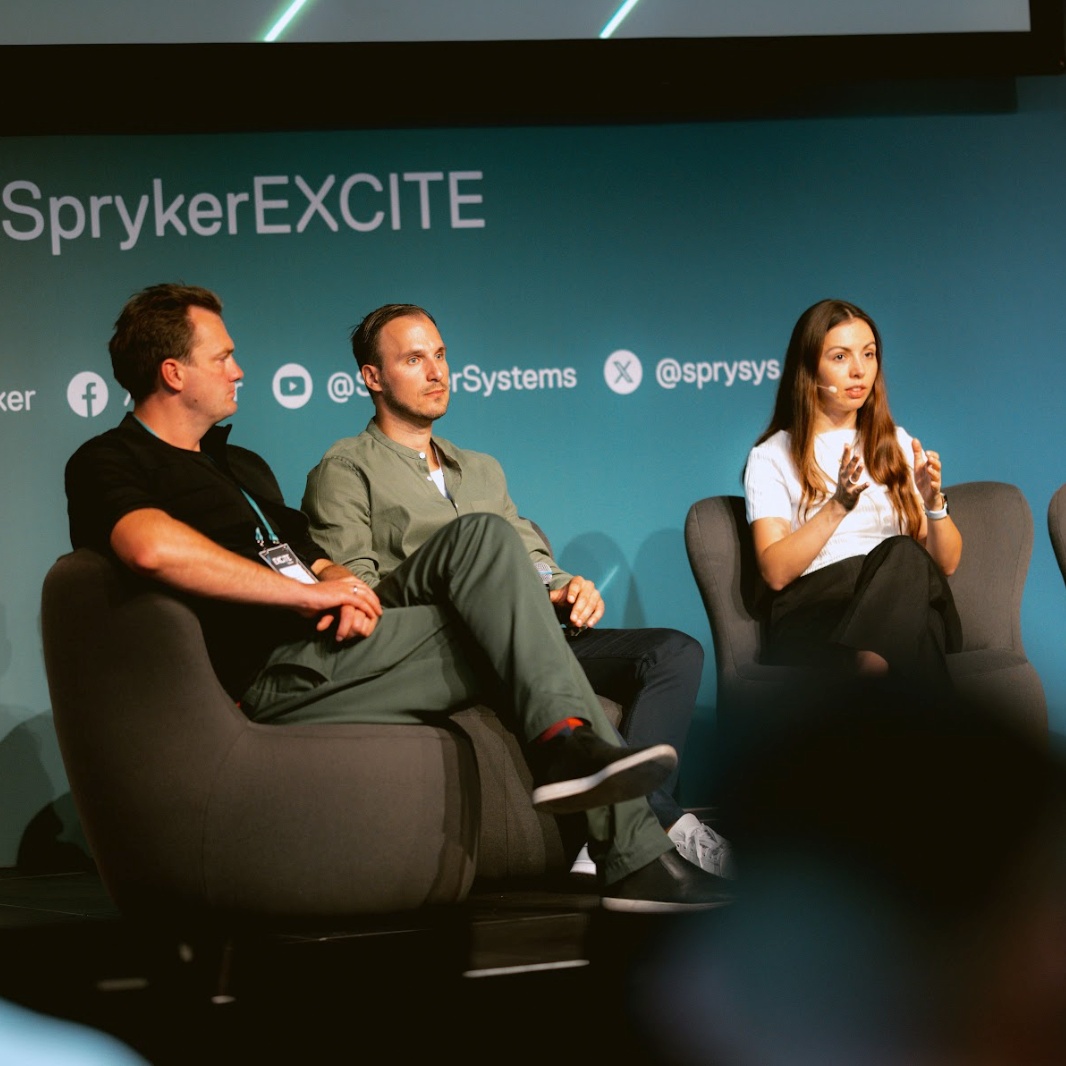 Three people seated on a stage in front of a blue backdrop with the text "Spryker EXCITE 2024." Two men on the left and a woman on the right are engaged in a discussion.