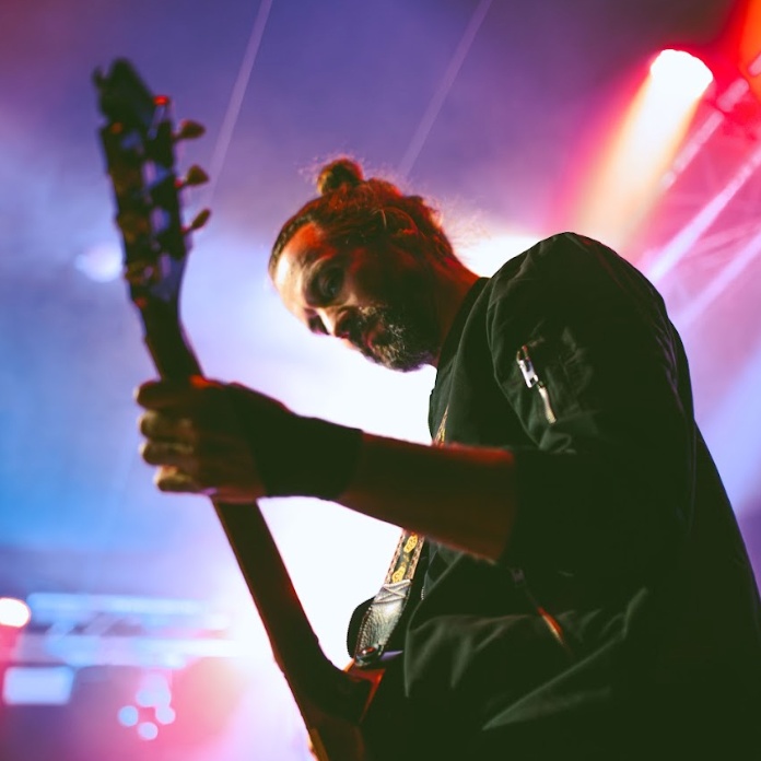 A musician plays an electric guitar on stage under colorful lighting at Spryker EXCITE 2024, wearing a dark long-sleeved shirt with his hair tied in a top knot.