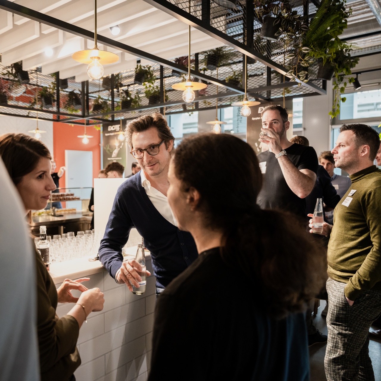 A group of people are socializing at an indoor event. They are gathered around a white countertop bar with drinks. The background has hanging plants, warm lighting, and a few posters. Everyone appears engaged in conversation.