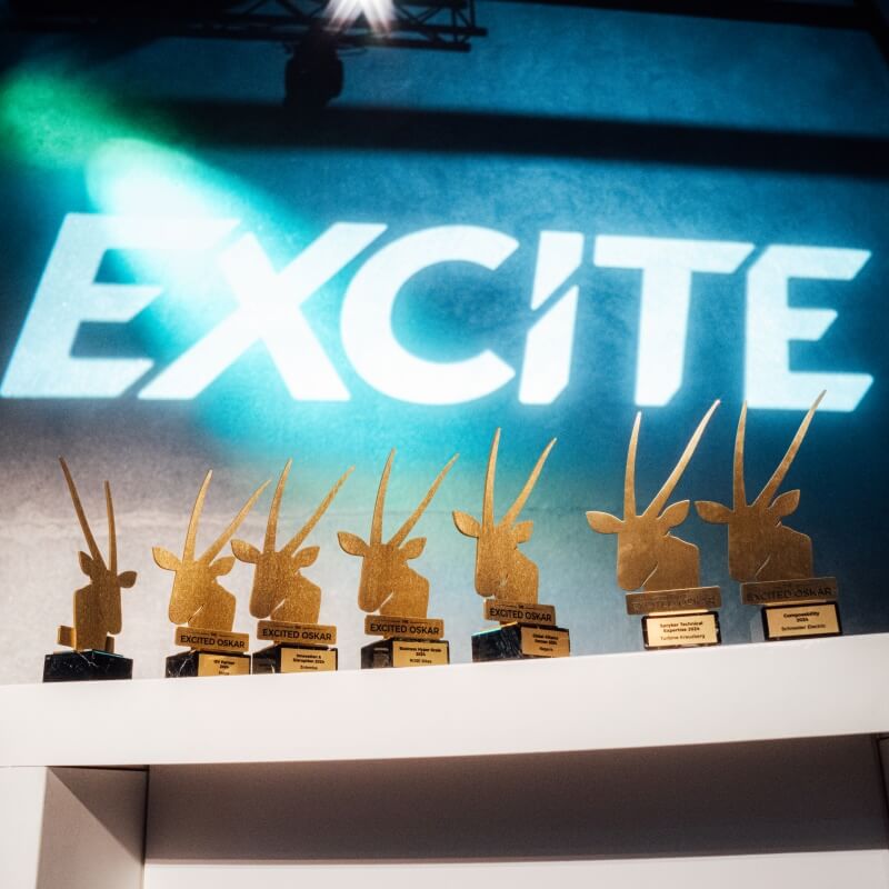 A row of gold antelope-shaped trophies is displayed on a white shelf, with "EXCITE" and the Spryker EXCITE 2025 branding illuminated on the wall behind them.