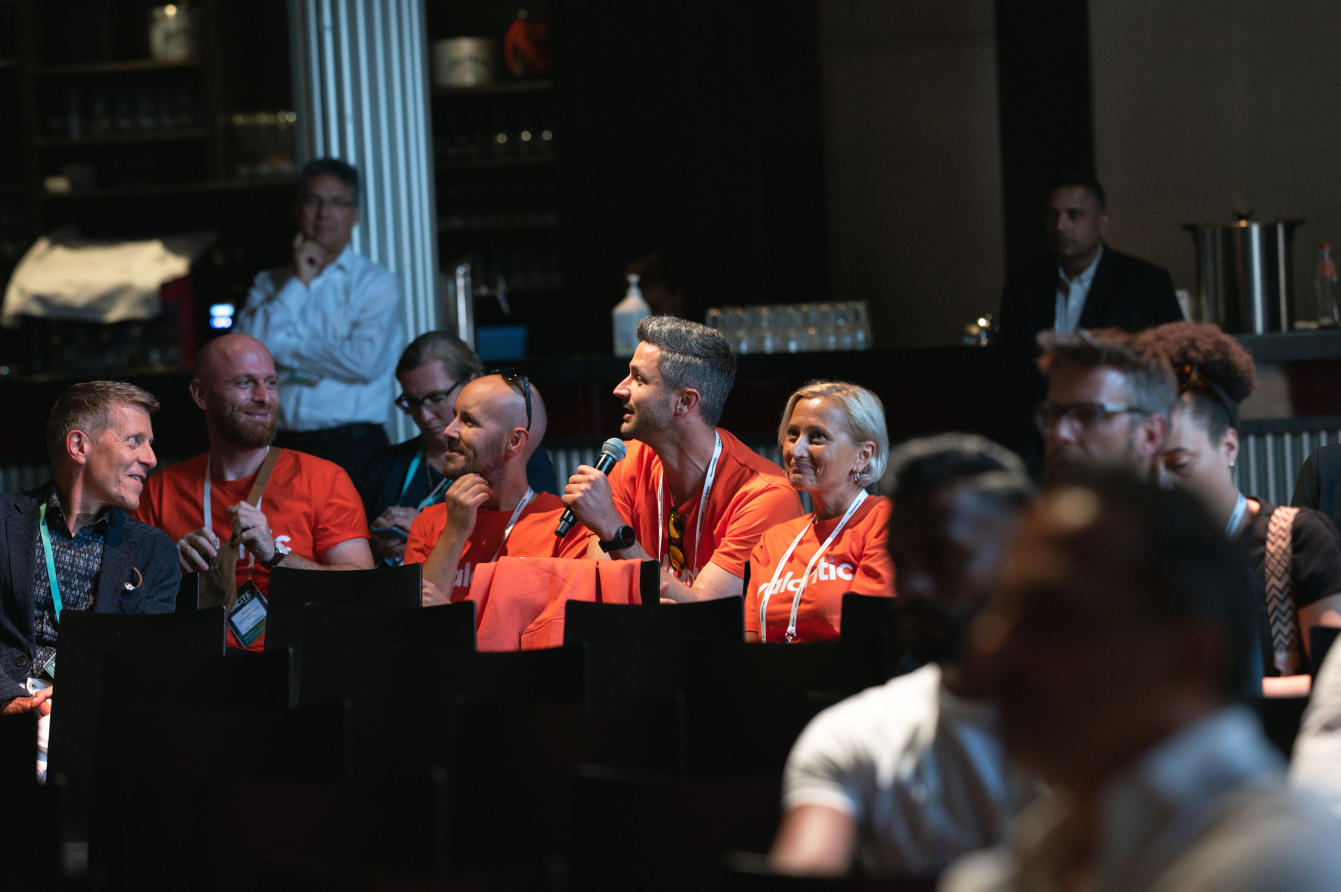 A group of people, some wearing orange shirts, are seated in a conference room at Spryker EXCITE 2024. One person is speaking into a microphone. Others are looking towards the speaker, while some are engaged in conversation.