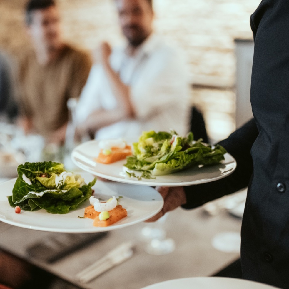 A waiter serves two plates of food, each featuring a lush leafy green salad, to a table with three people in the background, reminiscent of the vibrant atmosphere at Spryker EXCITE 2024.