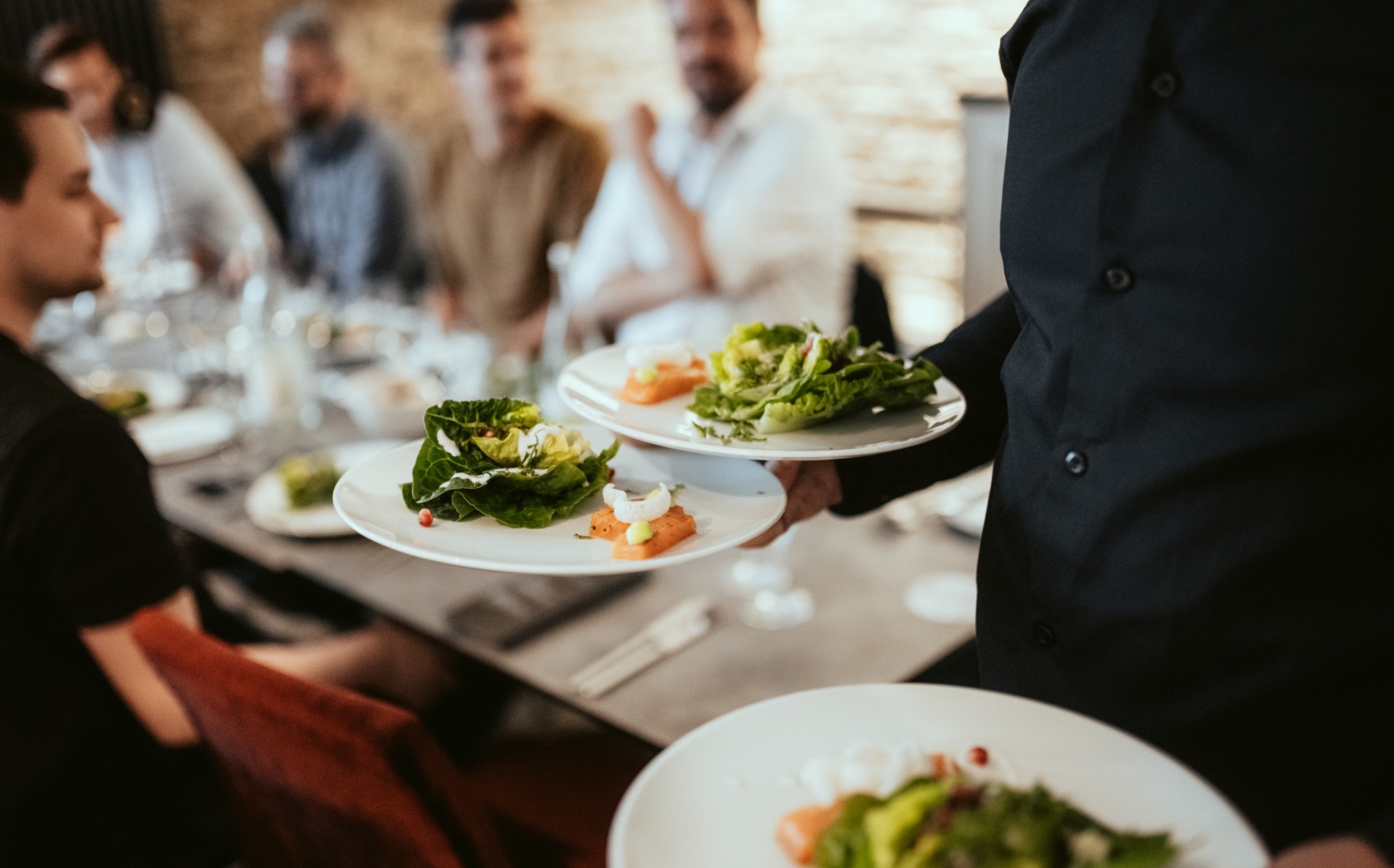 A server carries plates of salad while a group of people, buzzing with anticipation for Spryker EXCITE 2024, are seated at a dining table in the background.
