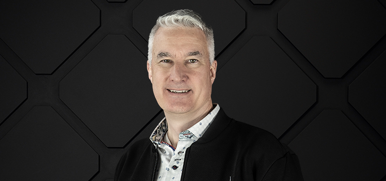 A man with short gray hair, exuding leadership in a dark collared shirt, stands in front of a black geometric background.