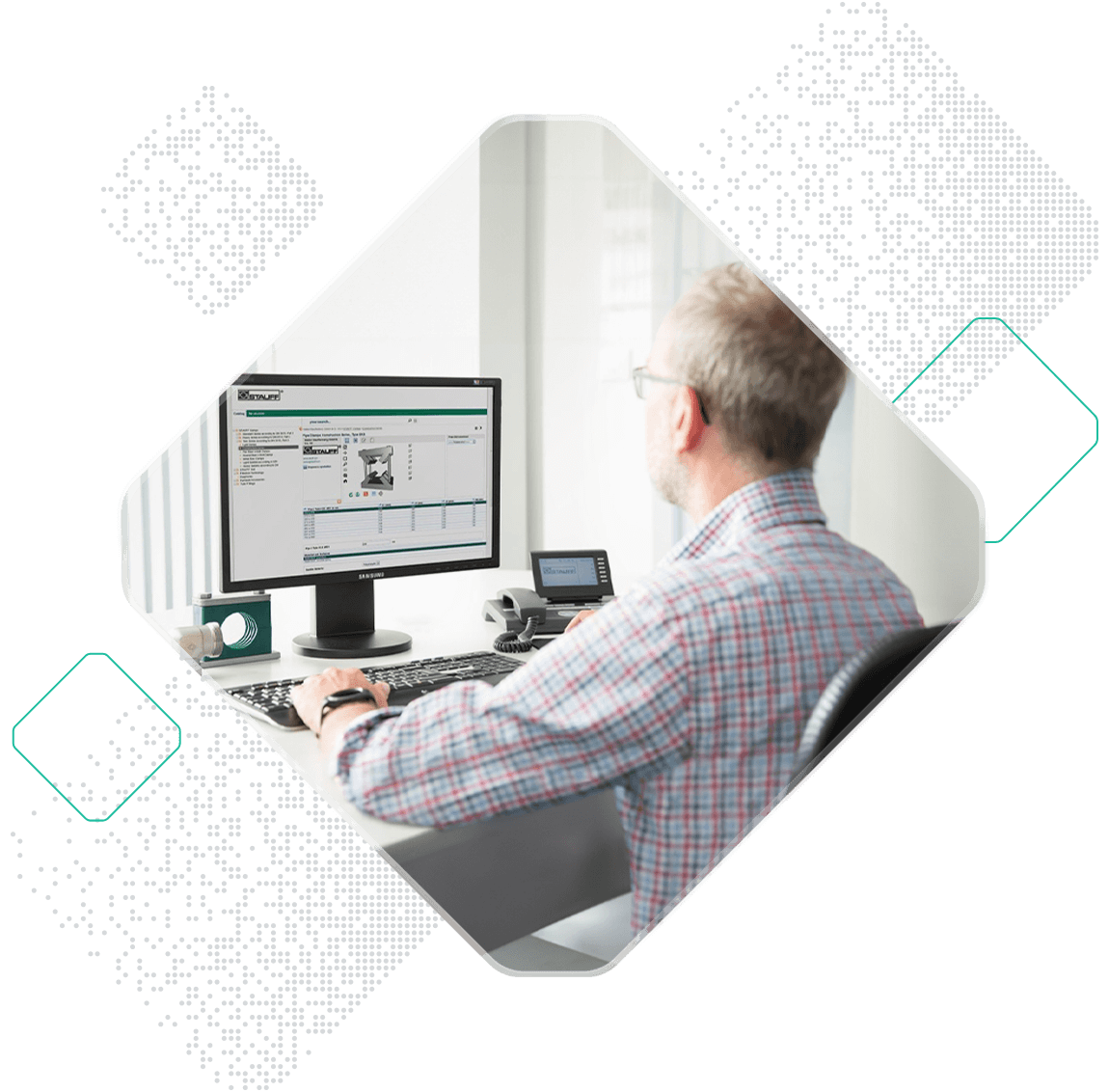 A person with gray hair and glasses, wearing a Stauff plaid shirt, is seated at a desk working on a computer. The computer screen displays a data management software interface.