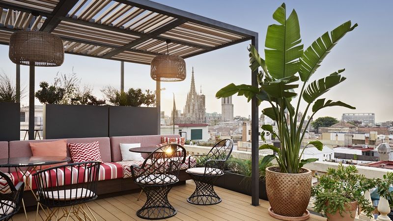 A cozy rooftop terrace featuring modern outdoor furniture, wicker chairs and a striped bench. A large potted plant stands beside the seating area under a pergola. The city skyline and a cathedral can be seen in the background.