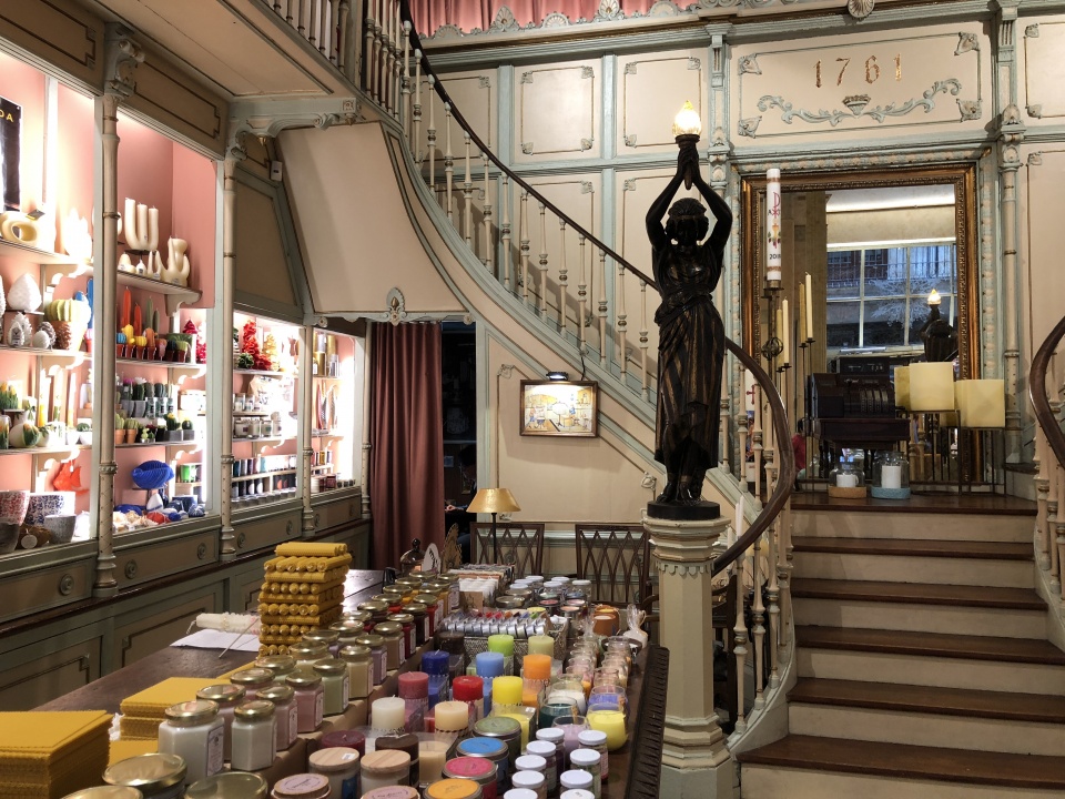 A quaint candle shop with a vintage interior features a wooden staircase and a variety of colorful candles and jars displayed on a table. Shelves on the left hold neatly arranged candles, while a statue holding a light stands by the stairs.