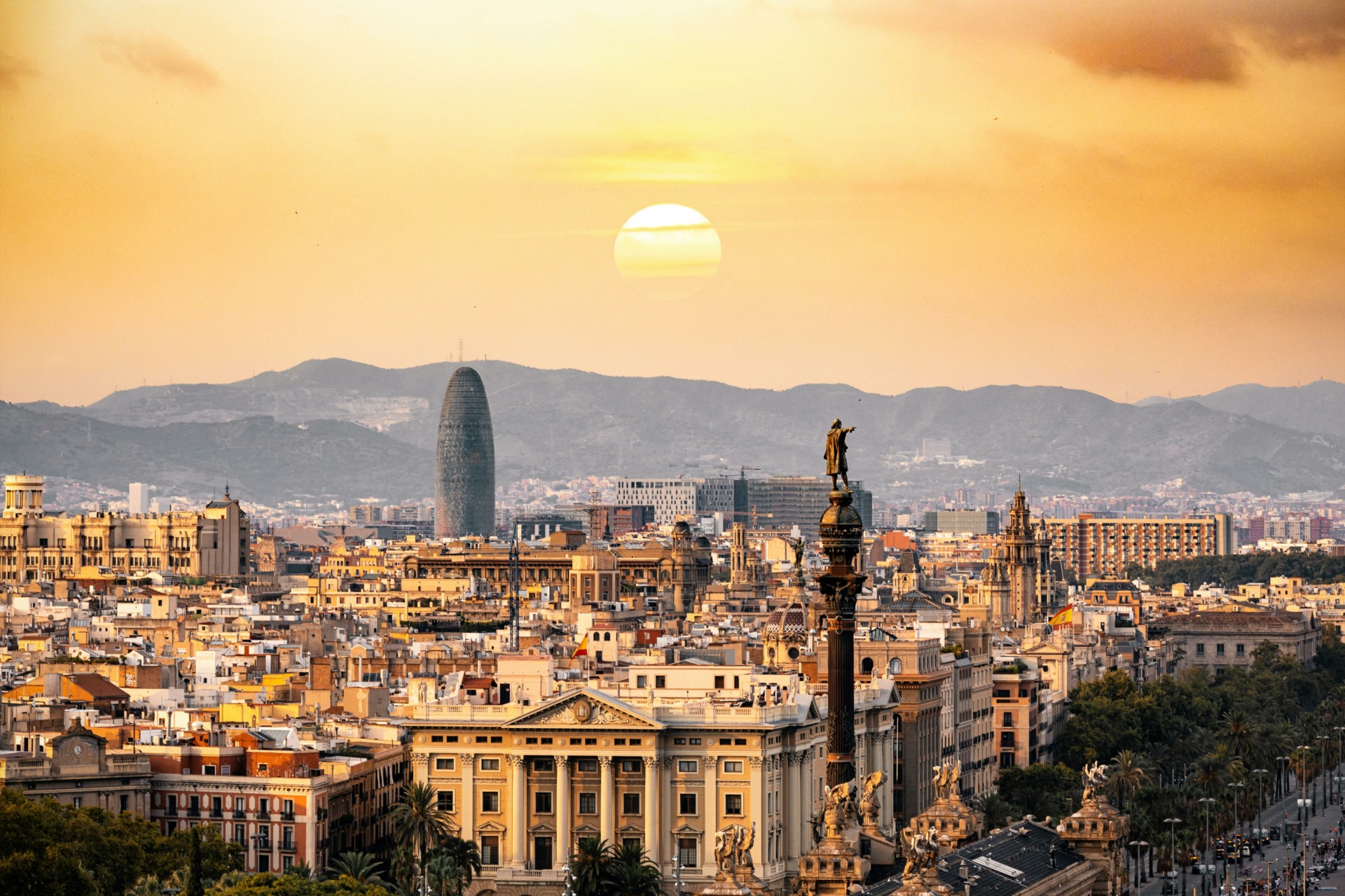 A cityscape of Barcelona at sunset, showcasing historical and modern architecture, with the Columbus Monument in the foreground and the distinctive, cylindrical Torre Glòries building in the background against a backdrop of mountains.