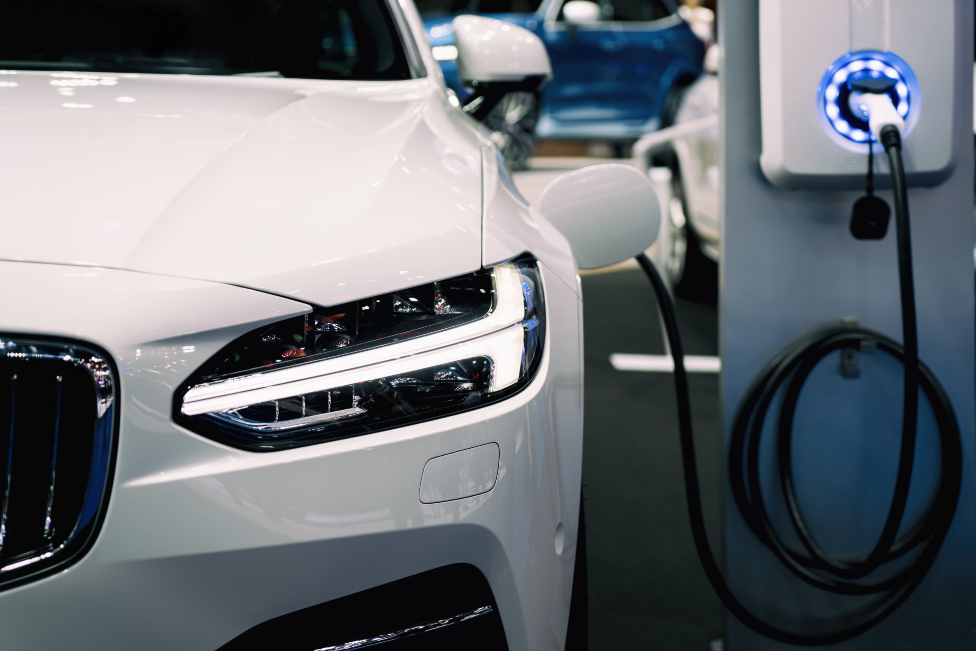 Close-up of a white electric car plugged into a charging station. The front headlight and part of the grille are visible, with the charging cable connected to the car. In the background, other cars are blurred. The scene depicts a modern electric vehicle charging setup.