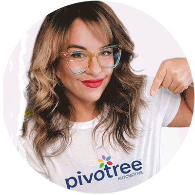 A person with wavy brown hair and colorful glasses smiles and points to their white shirt, which has the word "Pivotree" and "Automotive" printed on it along with a colorful logo.